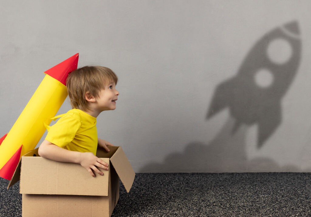 Small child wears a makeshift rocket backpack sitting in a box casting a shadow of a rocket launching.